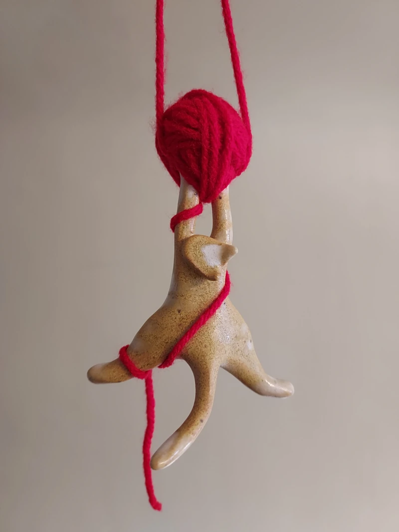 A playful tan ceramic hanging sculpture holding bright red yarn, in its original photograph